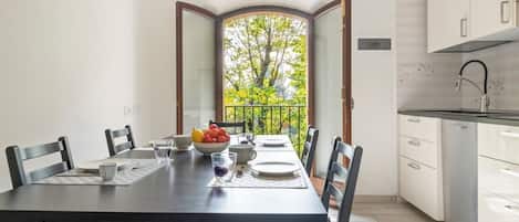 Un comedor con mesa dispuesta para cuatro, una cocina con armarios blancos y vista a árboles desde una puerta abierta.