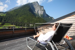 A person relaxing on a deck chair with a mountain view, a wooden deck, and a plate of apples. - Standard room - Hotel Krone Lingg GmbH & Co KG (Au)