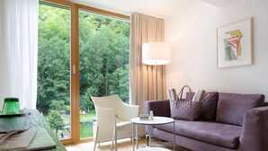 A modern living room with a sofa, a table, a lamp, and a view of a forest through large windows.