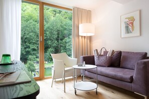 A modern living room with a sofa, a table, a lamp, and a view of a forest through large windows. - Standard room - Hotel Krone Lingg GmbH & Co KG (Au)