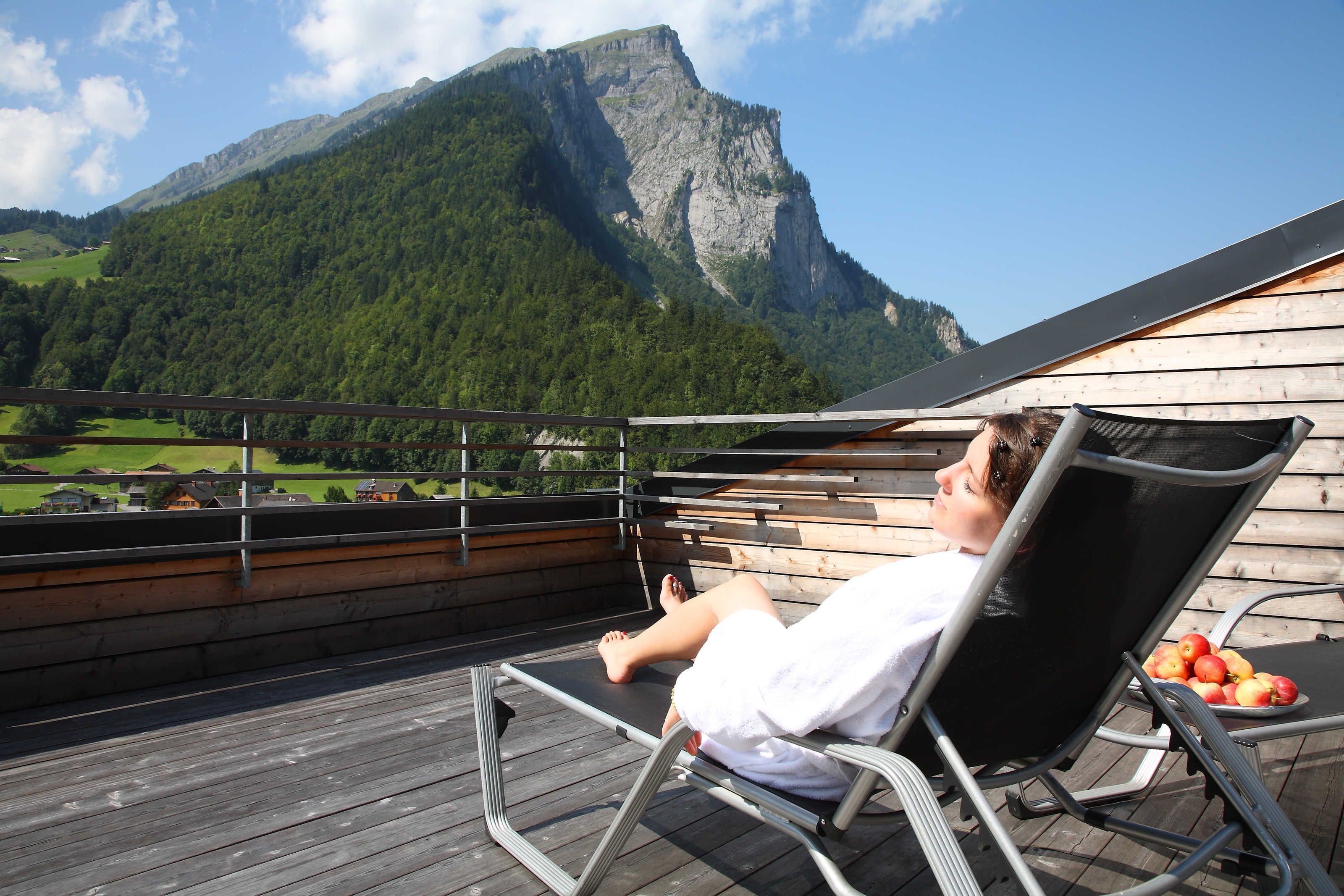 Eine Person entspannt sich in einer Liege mit Blick auf die Berge, einer Holzterrasse und einem Teller Äpfel.