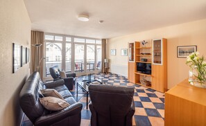 Ein Wohnzimmer mit einer Couch, Sesseln, einem Fernseher und einem Bücherregal.