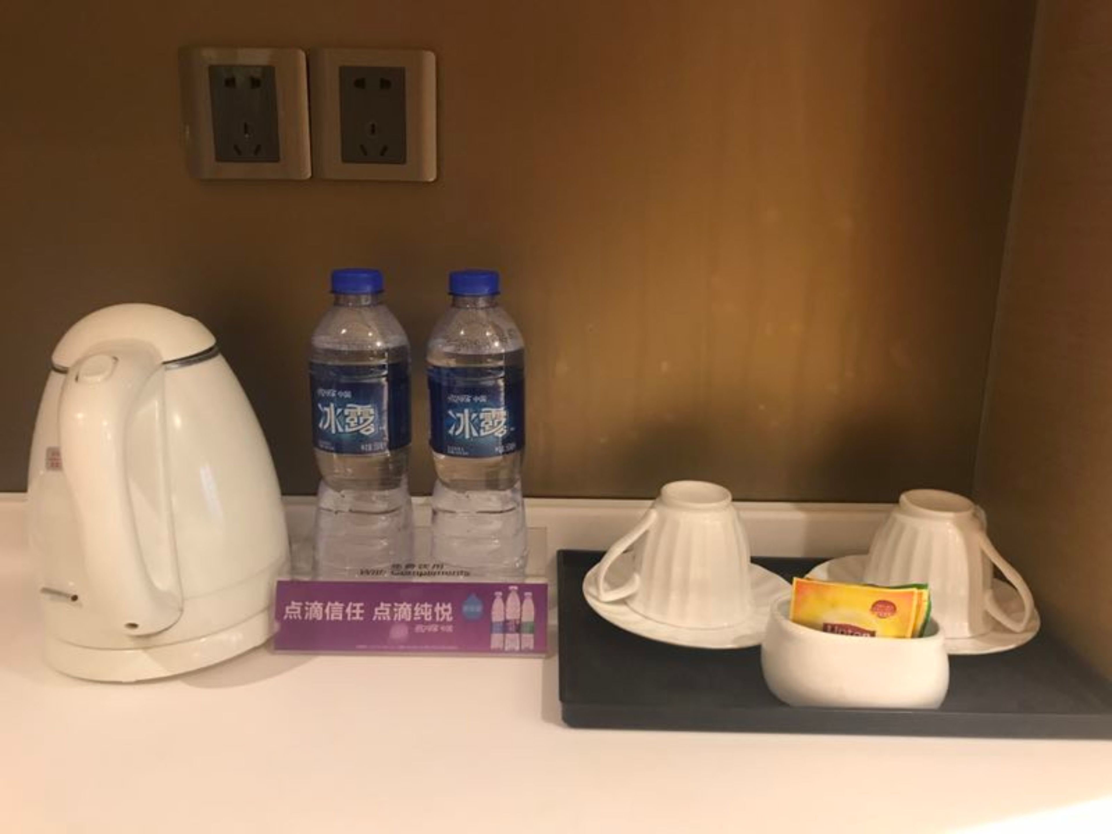 A hotel room with a white kettle, two bottles of water, a purple box, and two white cups on a tray.