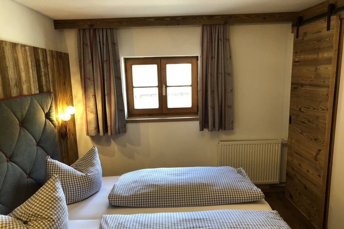 Un dormitorio con dos camas, ropa de cama a cuadros, una ventana con cortinas, una lámpara de pared y paneles de madera.