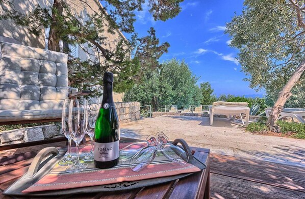 Une table avec une bouteille de vin, des verres et un set de table, le tout installé à l’extérieur, dans un décor composé de murs en pierre et d’arbres.