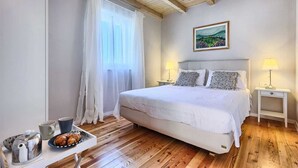 A bedroom with a bed, two bedside tables with lamps, a window with sheer curtains, a painting on the wall, and a tray with a teapot and croissants on the floor.