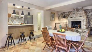 A dining area with a stone fireplace, wooden chairs, a round table, and a kitchen with modern appliances and pendant lights.