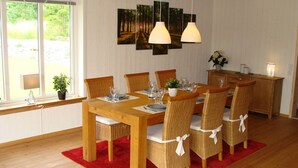 Un coin repas avec une table en bois dressée pour six personnes, entourée de chaises en osier ornées de rubans blancs. Trois lampes suspendues sont placées au-dessus de la table, et une fenêtre offre une vue sur la verdure à l’extérieur.