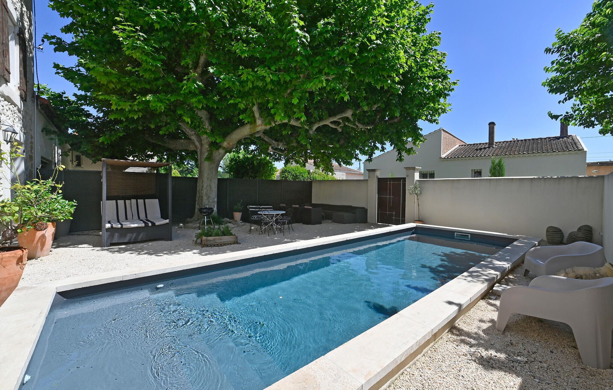 A rectangular swimming pool with a tiled deck, surrounded by a gravel area. There is a wooden bench and a table with chairs under a tree.
