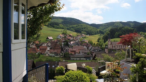 Blick auf ein Dorf mit Häusern und einer Kirche, umgeben von Hügeln und Grünflächen.