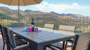 A terrace with a table, chairs, and an umbrella. A bottle of wine is on the table. The terrace overlooks a mountainous landscape. - Stunning home in Nessa (Nessa)