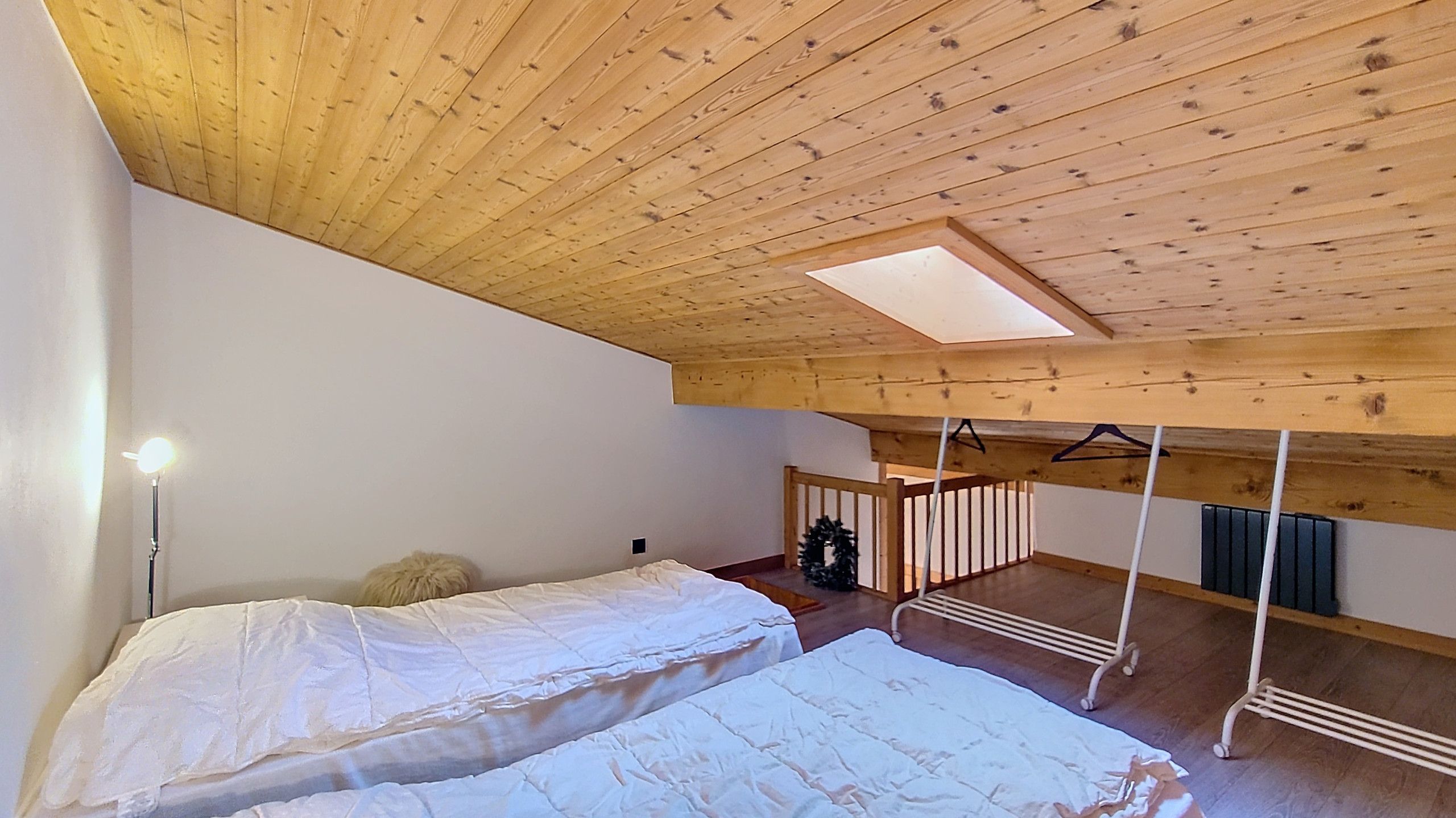 A room with a sloped ceiling, wooden beams, a bed with white bedding, a hanging light fixture, and a small Christmas wreath.