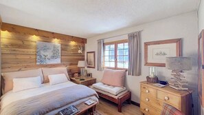A cozy bedroom with a wooden headboard, a large bed, a bedside table with a lamp, a small chair, a dresser, and a window with curtains. - 1 Golf Aval (Megève)
