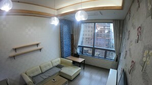 A modern living room with a sofa, a coffee table, a TV, and a standing fan. There are wooden shelves on the wall and a window with a view of buildings. - Gangnam Residence 4 (Seoul)
