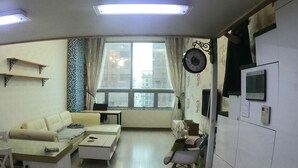 A compact living space with a sofa, a small table, a chair, a window with curtains, and a wall-mounted TV. - Gangnam Residence 4 (Seoul)