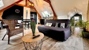 A cozy loft with a high ceiling, exposed wooden beams, a black sofa with patterned cushions, a wicker chair, a small round table with a vase and remote, and a kitchen in the background. - La Rose des Vents 4 people  (Grandcamp-Maisy)