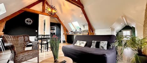 A cozy loft with a high ceiling, exposed wooden beams, a black sofa with patterned cushions, a wicker chair, a small round table with a vase and remote, and a kitchen in the background.