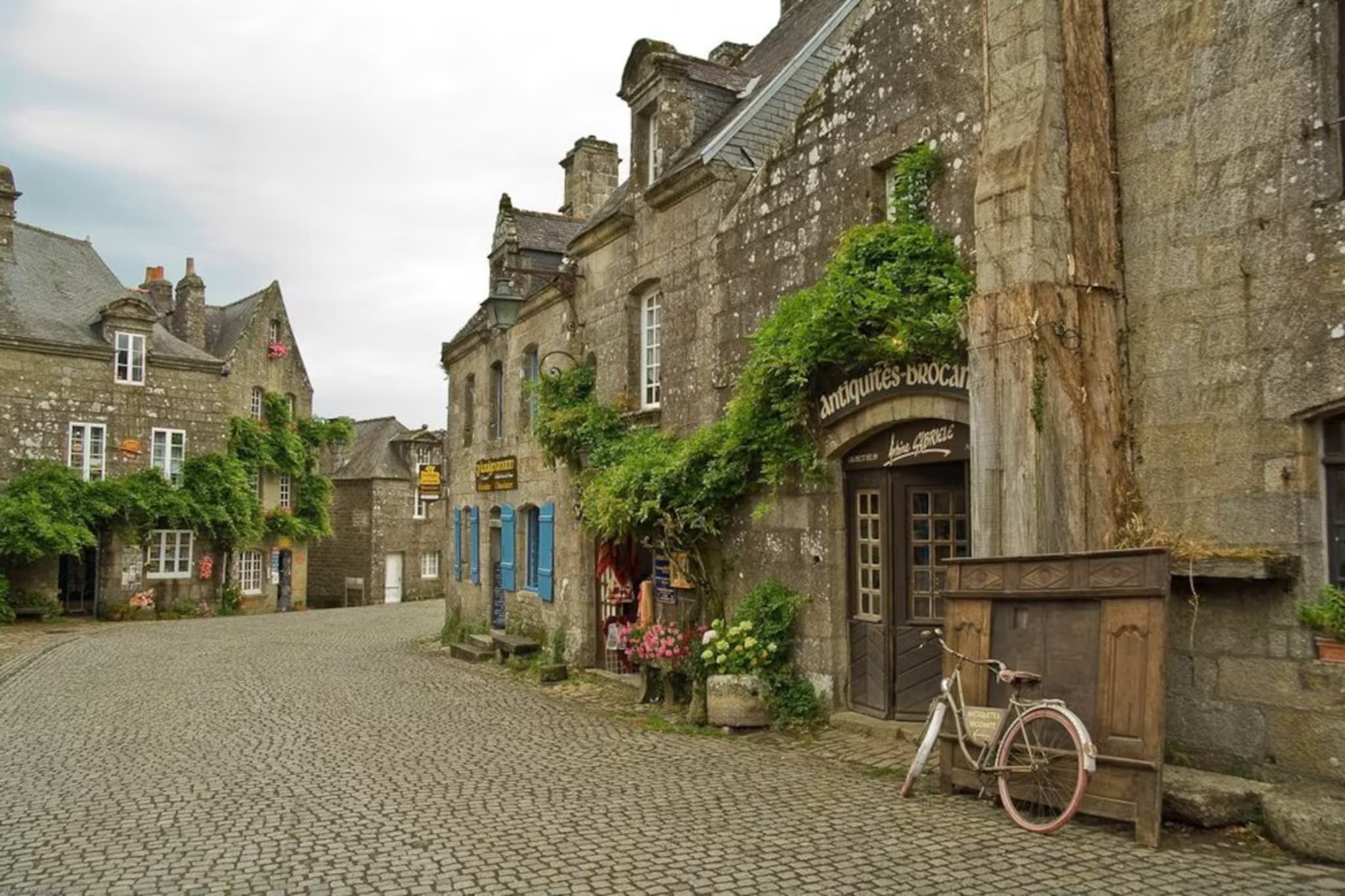 A cobblestone street lined with historic stone buildings, some with ivy and signage indicating antique shops.