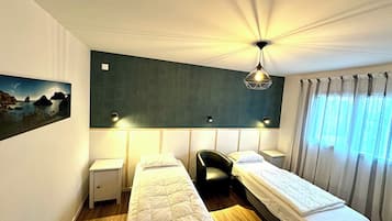 A hotel room with two single beds, a desk, a chair, a nightstand, a wall-mounted lamp, and a painting of a landscape.