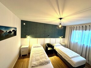 A hotel room with two single beds, a desk, a chair, a nightstand, a wall-mounted lamp, and a painting of a landscape.