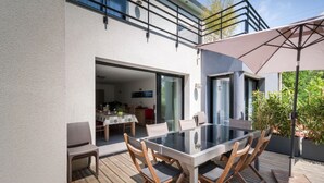En udendørs patio med et glasbord og træstole, et overdækket siddeområde og udsigt til indretningen indeni.