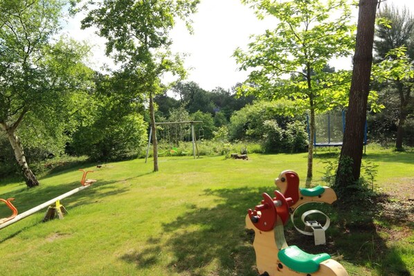 Un parque infantil con columpios y un sube y baja en un área verde rodeada de árboles.
