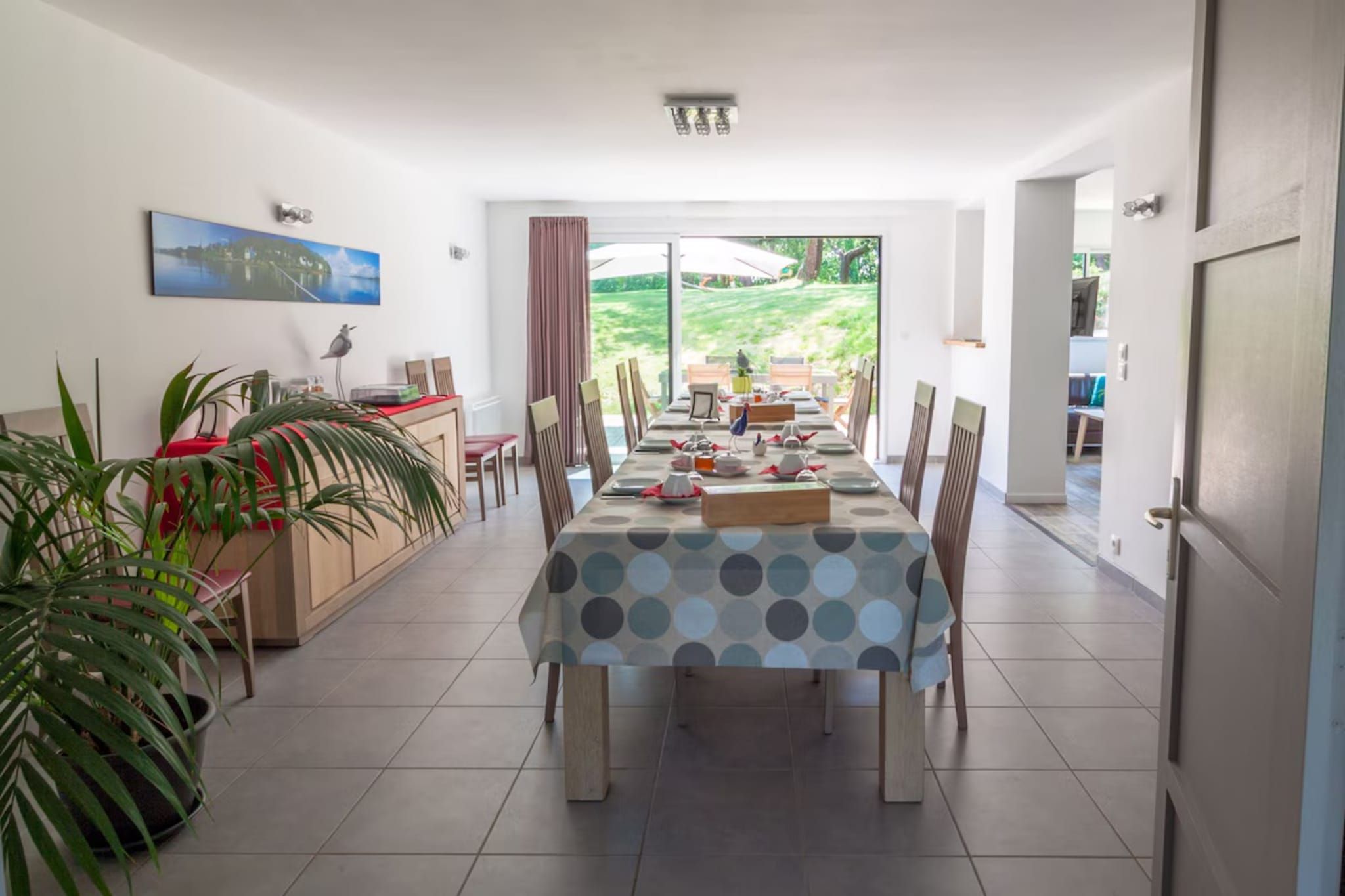 Un coin repas avec une table dressée pour quatre personnes, un espace cuisine avec un comptoir et des chaises, et une grande fenêtre donnant sur un paysage verdoyant.