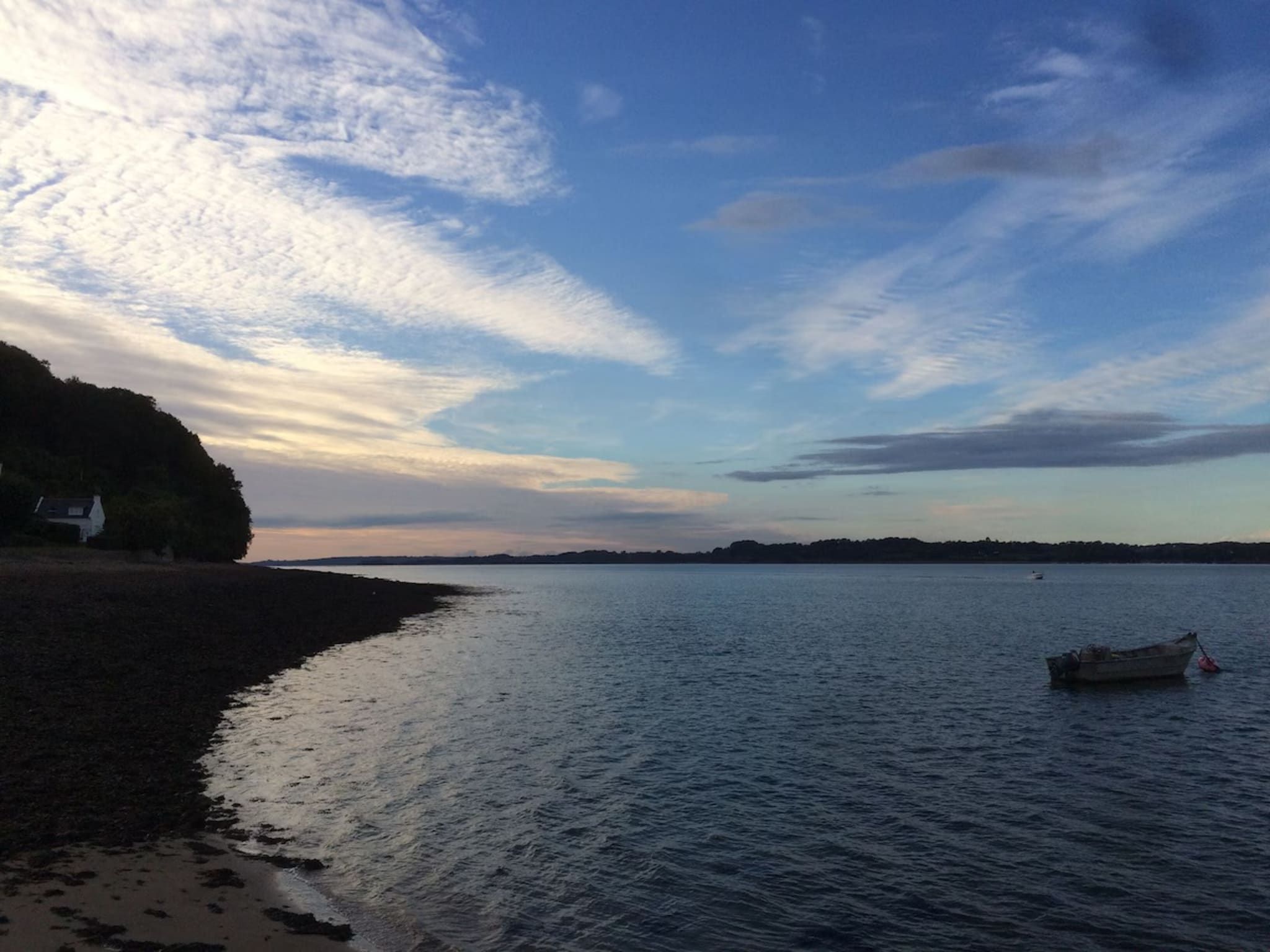 Une plage bordée d’une mer calme, un bateau sur l’eau et une côte lointaine.