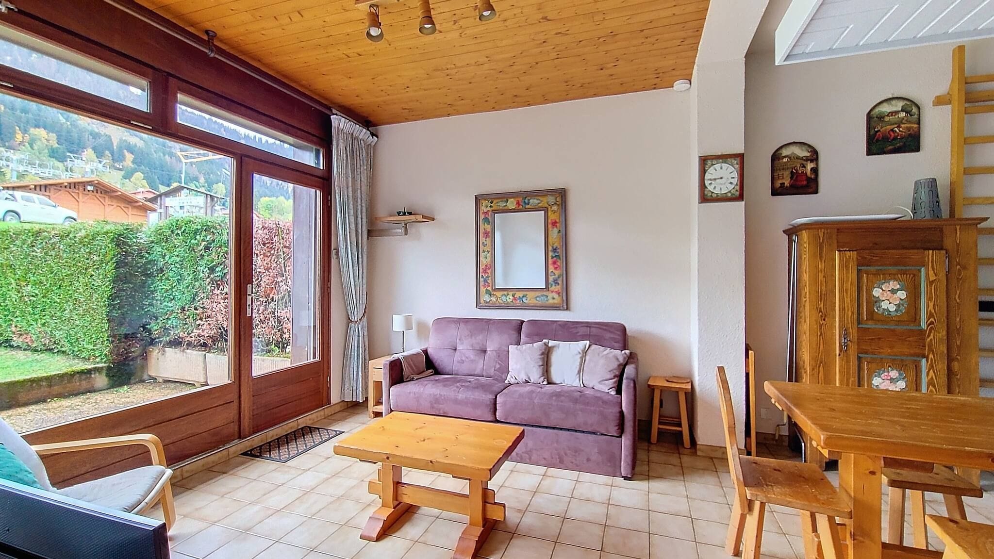 Un salon avec un canapé, une table en bois et une vue sur un jardin, visible à travers de grandes fenêtres.