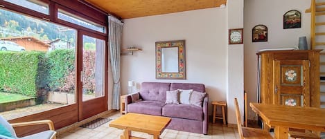 A living room with a sofa, a wooden table, and a view of a garden through large windows.