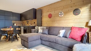 A modern living room with a grey sectional sofa, a dining table with chairs, and a kitchen area in the background.