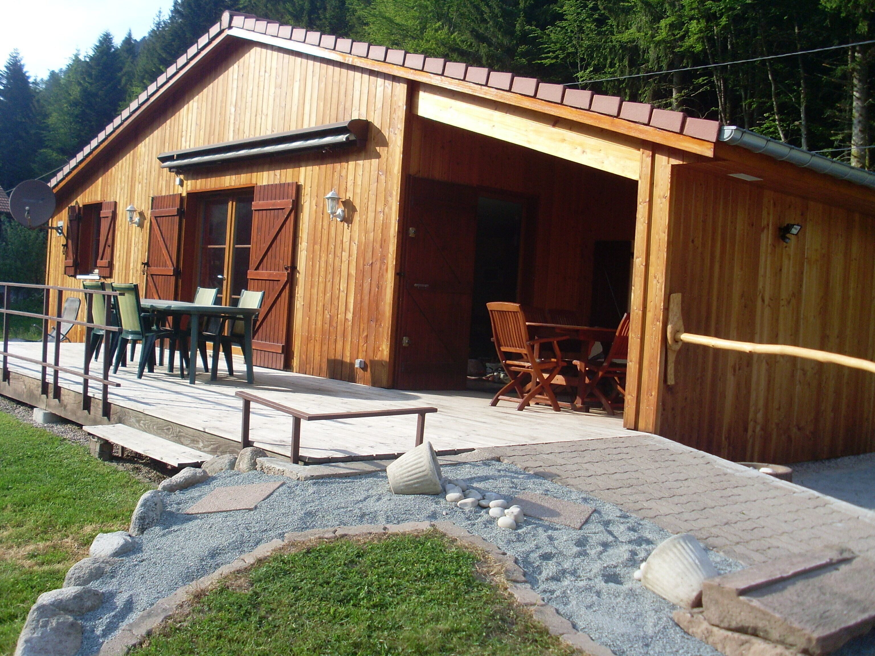 Een houten hut met een overdekt terras, buitenmeubels en een grindpad dat naar de ingang leidt.