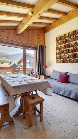 Una habitación con techo de madera, un ventanal con cortinas, un sofá con cojines, una mesa con mantel y una pared decorada con un tapiz.