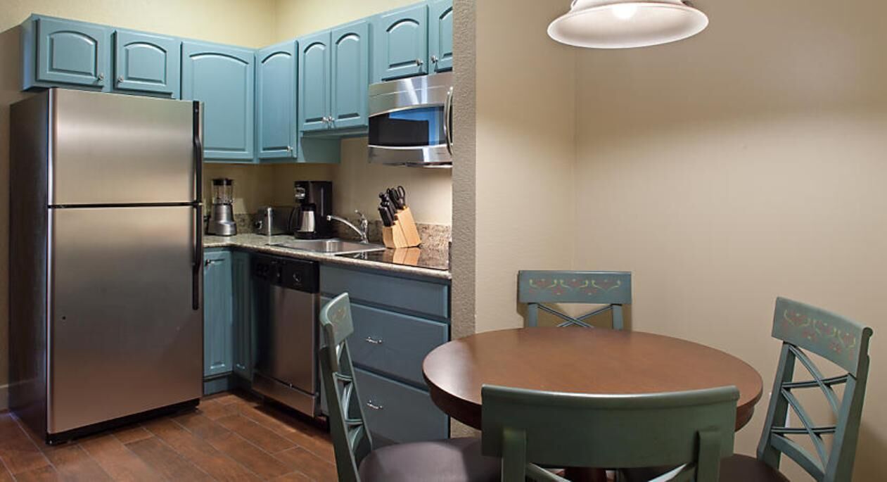 Cocina compacta con armarios azul verdoso, un refrigerador de acero inoxidable, un microondas y un lavabo. Hay una mesa redonda de madera con sillas.