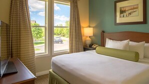Ein Hotelzimmer mit einem Bett, einem Schreibtisch, einer Lampe, einem gerahmten Bild an der Wand und einem Fenster mit Vorhängen.