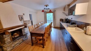 A kitchen with a wooden table and chairs, a stone fireplace, and a window with curtains.