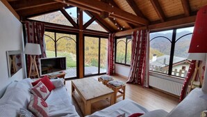 A cozy living room with a sofa, a wooden coffee table, and a TV. Large windows offer a view of a mountain landscape.