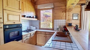 A compact kitchen with wooden cabinets, a stove, a sink, and a window offering a view of the outdoors. - 1 Chalet Clarines (Megève)