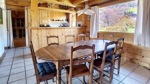 A wooden cabin interior with a dining table and chairs, a kitchen area with wooden cabinets, and a large window offering a view of the outdoors. - 1 Chalet Clarines (Megève)