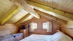 A cozy attic bedroom with a wooden ceiling, a bed, a dresser, a television, and two windows with curtains. - 1 Chalet Clarines (Megève)
