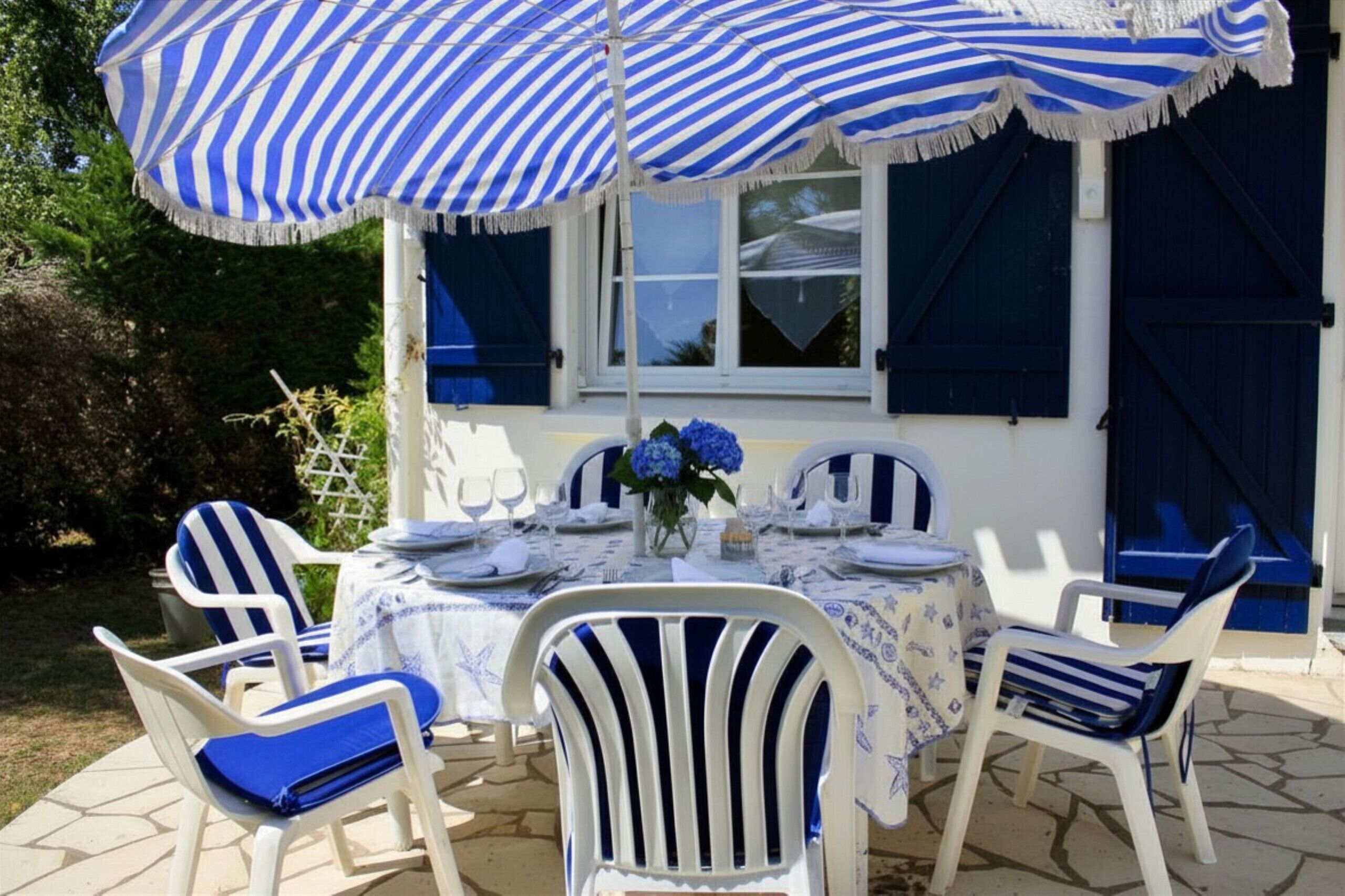Une terrasse avec des chaises blanches et des coussins bleus, une table dressée pour le repas et un parasol à rayures.