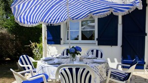 Een terras met witte stoelen en blauwe kussens, een gedekte tafel en een gestreept zonnescherm.