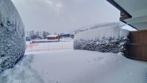 Un espace extérieur enneigé, avec une clôture en bois, un buisson recouvert de neige et un bâtiment en arrière-plan.