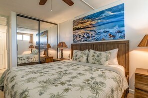 A bedroom with a large bed, a wooden headboard, a painting of a beach scene, and a mirror reflecting the room.