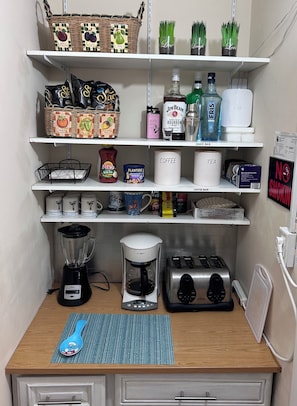Una kitchenette con cafetera, tostadora, licuadora y diferentes alimentos en estantes.