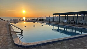 Ein Schwimmbecken mit einer gefliesten Kante, ein überdachter Sitzbereich mit Liegestühlen und Blick auf den Sonnenuntergang über dem Wasser.
