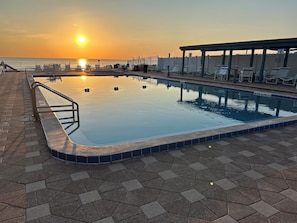 Uma piscina com borda revestida em azulejo, área de estar coberta com espreguiçadeiras e vista do pôr do sol sobre a água.