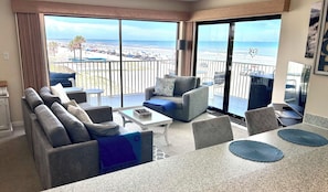 Habitación de hotel con balcón, un conjunto de sofás, una mesa de comedor y vistas a la playa.