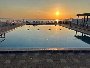 A swimming pool with a view of the sunset over the ocean, surrounded by lounge chairs and a fence.