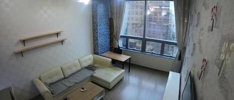 A modern living room with a sofa, a coffee table, a TV, and a standing fan. There are wooden shelves on the wall and a window with a view of buildings.